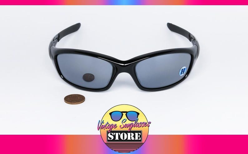 May include: Black sunglasses with gray lenses. The sunglasses have a curved frame and a small sticker on one lens. A penny is next to the sunglasses. The image also includes a logo for a vintage sunglasses store.