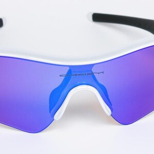 Pu&ograve; includere: Occhiali da sole sportivi bianchi con lenti a specchio viola e arancioni. Gli occhiali hanno una montatura bianca con dettagli neri sulle aste. Il marchio "Oakley" &egrave; visibile sulla lente.