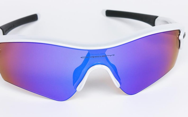 Pu&ograve; includere: Occhiali da sole sportivi bianchi con lenti a specchio viola e arancioni. Gli occhiali hanno una montatura bianca con dettagli neri sulle aste. Il marchio "Oakley" &egrave; visibile sulla lente.