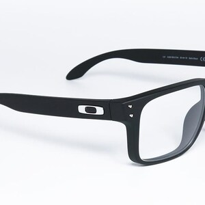 May include: Black rectangular eyeglasses with clear lenses. The frames are matte black with a silver logo on the temples. The glasses have a modern, sporty design, suitable for everyday wear.