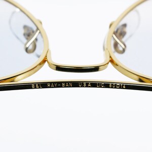 Op de afbeelding: Close-up van de inscriptie op de poot van een paar gouden aviator-zonnebrillen. De inscriptie luidt "B&L RAY-BAN U.S.A. LIC. 60014".