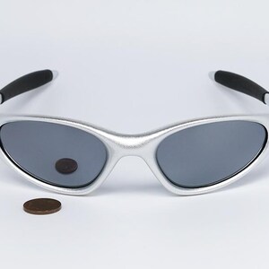 May include: Silver-framed sunglasses with dark gray lenses and black temple tips. A penny is next to the sunglasses. The sunglasses have a sleek, modern design, suitable for outdoor activities.