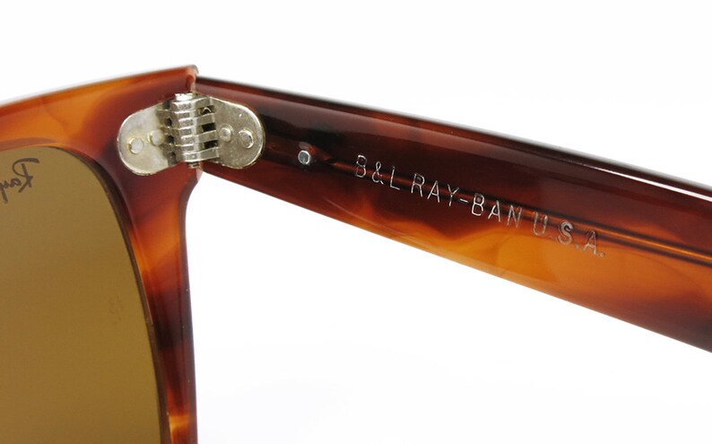 Peut inclure: Gros plan sur la charni&egrave;re et la branche d'une paire de lunettes de soleil Ray-Ban en &eacute;caille de tortue brune. La branche est grav&eacute;e "B&L RAY-BAN U.S.A."