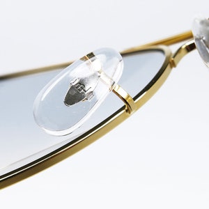 Op de afbeelding: Close-up van de gouden metalen frame van een zonnebril met transparante kunststof oorsteunen. De oorsteunen hebben een klein zilveren logo.