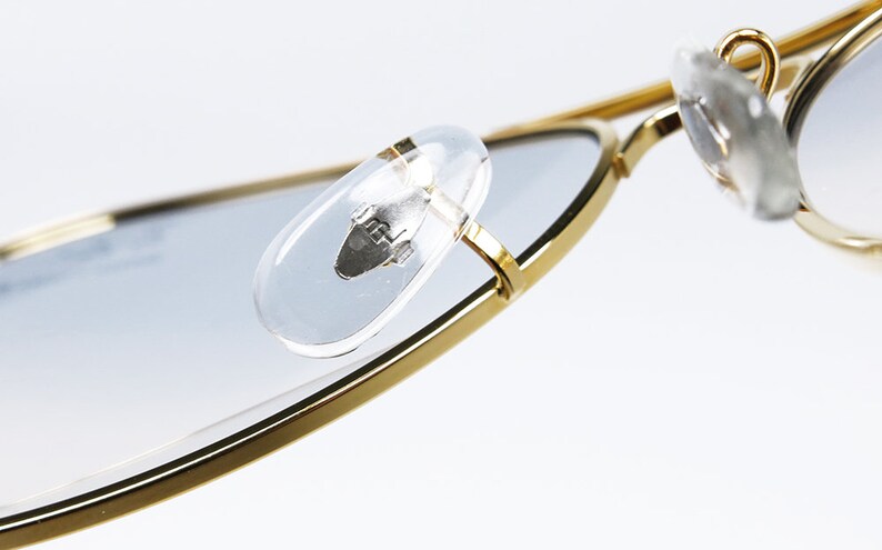 Op de afbeelding: Close-up van de gouden metalen frame van een zonnebril met transparante kunststof oorsteunen. De oorsteunen hebben een klein zilveren logo.