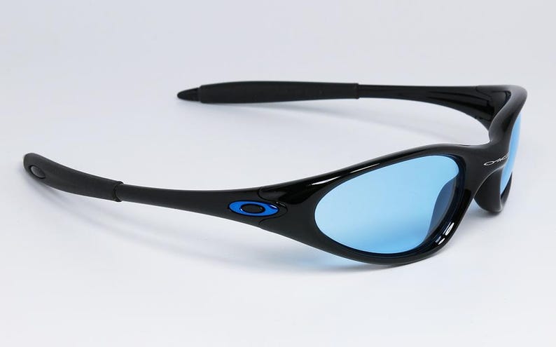 May include: Black sunglasses with blue lenses. The sunglasses have a sleek, curved design with black frames and temples. A small blue accent is visible on the frame near the temple. The lenses are a light blue color.