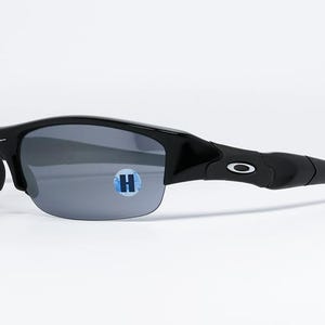 K&ouml;nnte beinhalten: Schwarze Sonnenbrille mit grauen Gl&auml;sern. Die Sonnenbrille hat ein geschwungenes Design und ein kleines wei&szlig;es Logo am B&uuml;gel. Auf dem Glas befindet sich ein blau-wei&szlig;er Aufkleber.