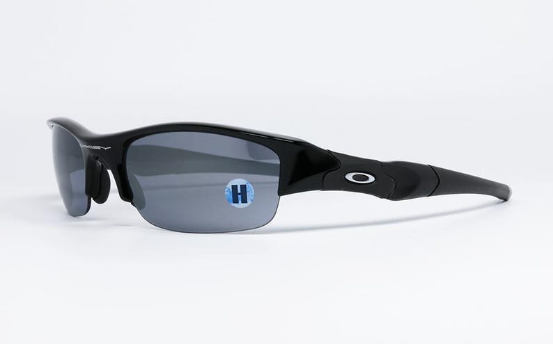 K&ouml;nnte beinhalten: Schwarze Sonnenbrille mit grauen Gl&auml;sern. Die Sonnenbrille hat ein geschwungenes Design und ein kleines wei&szlig;es Logo am B&uuml;gel. Auf dem Glas befindet sich ein blau-wei&szlig;er Aufkleber.