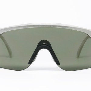 Puede incluir: Un par de gafas de sol con montura plateada y lentes verde oscuro. Las gafas tienen un dise&ntilde;o angular y futurista.