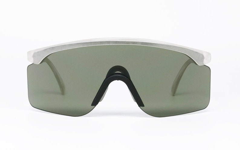 Puede incluir: Un par de gafas de sol con montura plateada y lentes verde oscuro. Las gafas tienen un dise&ntilde;o angular y futurista.