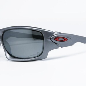 May include: Gray sunglasses with dark lenses. The sunglasses have a wraparound design with a red and black logo on the temple. The lenses have a slight curve and the frame is a matte gray color. The sunglasses are designed for sports and outdoor activities.