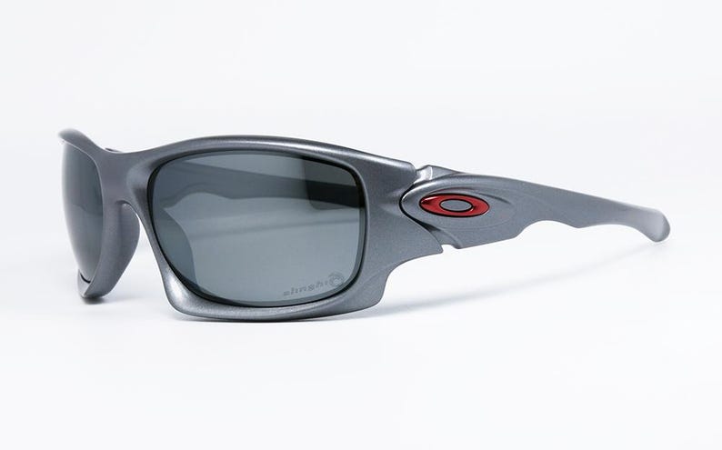 May include: Gray sunglasses with dark lenses. The sunglasses have a wraparound design with a red and black logo on the temple. The lenses have a slight curve and the frame is a matte gray color. The sunglasses are designed for sports and outdoor activities.