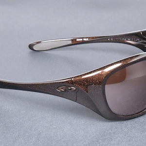 K&ouml;nnte beinhalten: Braune und schwarze Sonnenbrille mit Metallic-Finish. Die Sonnenbrille hat einen geschwungenen Rahmen und dunkle Gl&auml;ser. Das Oakley-Logo ist auf dem Rahmen sichtbar. Die B&uuml;gel der Sonnenbrille sind gebogen.