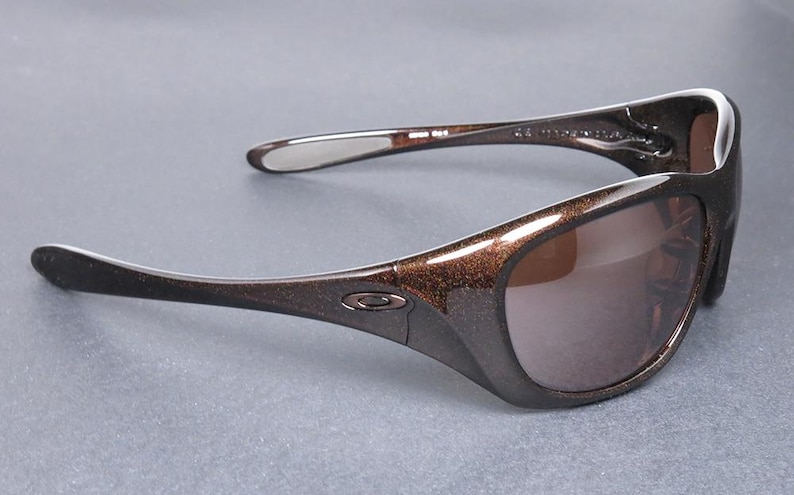 K&ouml;nnte beinhalten: Braune und schwarze Sonnenbrille mit Metallic-Finish. Die Sonnenbrille hat einen geschwungenen Rahmen und dunkle Gl&auml;ser. Das Oakley-Logo ist auf dem Rahmen sichtbar. Die B&uuml;gel der Sonnenbrille sind gebogen.