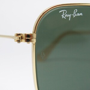 May include: Close-up of Ray-Ban sunglasses. The sunglasses have a gold-colored frame and green lenses. The Ray-Ban logo is visible on the upper left lens. The hinge and arm of the sunglasses are also visible.
