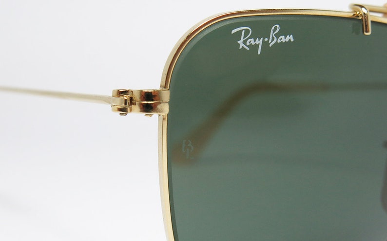 May include: Close-up of Ray-Ban sunglasses. The sunglasses have a gold-colored frame and green lenses. The Ray-Ban logo is visible on the upper left lens. The hinge and arm of the sunglasses are also visible.