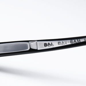 May include: Close-up of a black Ray-Ban eyeglass arm. The arm features a silver metal plate with the engraved text "B&L RAY-BAN U.S.A." A small screw secures the plate to the black frame. The background is white.