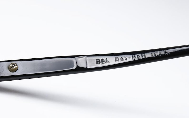 May include: Close-up of a black Ray-Ban eyeglass arm. The arm features a silver metal plate with the engraved text "B&L RAY-BAN U.S.A." A small screw secures the plate to the black frame. The background is white.