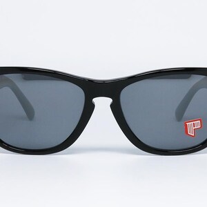 May include: Black sunglasses with gray lenses. The sunglasses have a thick, rectangular frame and a small red and white logo on the right lens. The sunglasses are set against a white background.