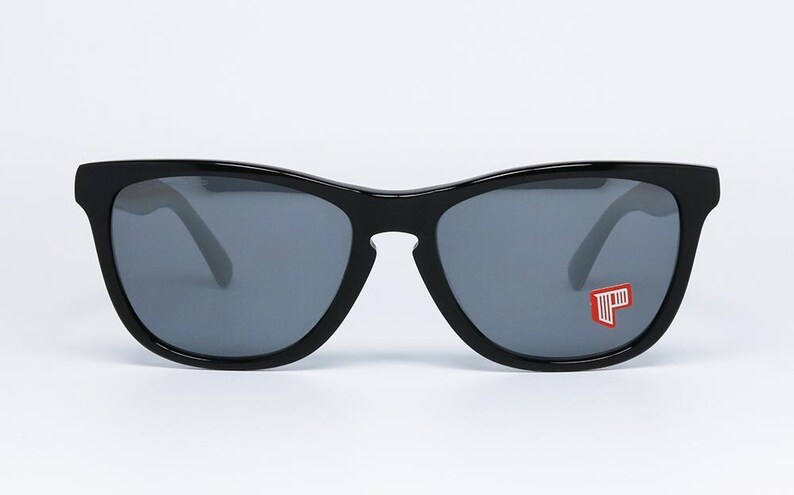 May include: Black sunglasses with gray lenses. The sunglasses have a thick, rectangular frame and a small red and white logo on the right lens. The sunglasses are set against a white background.