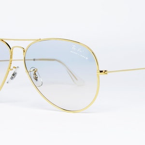 Op de afbeelding: Gouden aviator zonnebril met lichtblauwe gradient lenzen. De zonnebril heeft een Ray-Ban logo op de rechter lens.