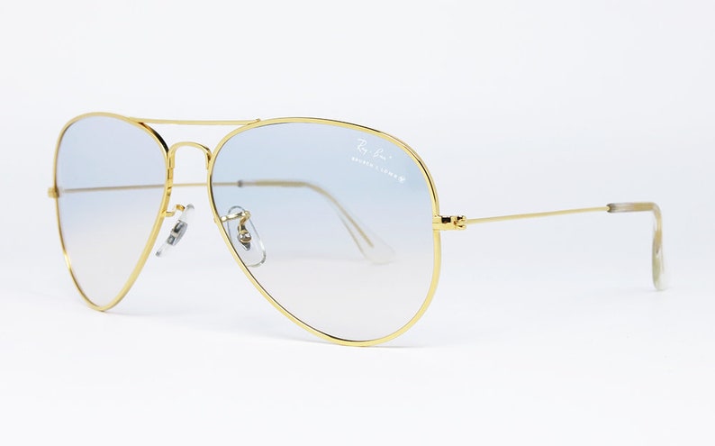 Op de afbeelding: Gouden aviator zonnebril met lichtblauwe gradient lenzen. De zonnebril heeft een Ray-Ban logo op de rechter lens.