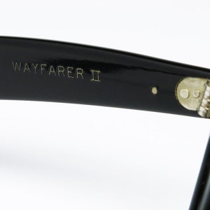 K&ouml;nnte beinhalten: Nahaufnahme des schwarzen Kunststoffgestells einer Wayfarer II Sonnenbrille. Die Sonnenbrille hat ein Metallgelenk und der Text "WAYFARER II" ist in das Gestell graviert.