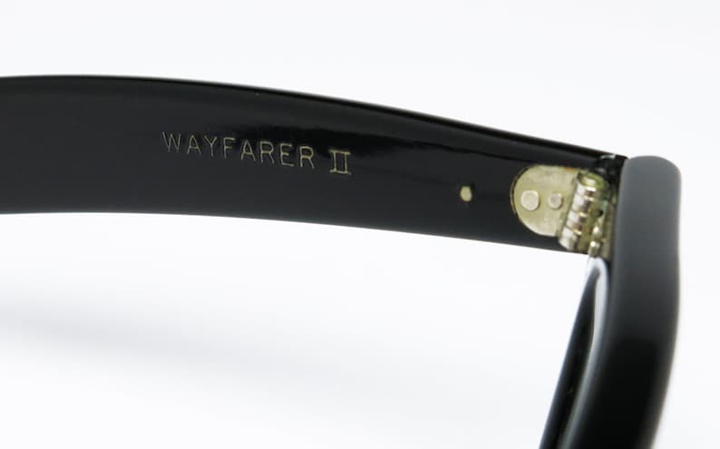 K&ouml;nnte beinhalten: Nahaufnahme des schwarzen Kunststoffgestells einer Wayfarer II Sonnenbrille. Die Sonnenbrille hat ein Metallgelenk und der Text "WAYFARER II" ist in das Gestell graviert.