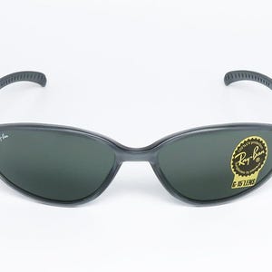 May include: Dark gray Ray-Ban sunglasses with dark green lenses. The sunglasses have a curved frame and a sticker on the right lens that reads "Ray-Ban G-15 LENS". The Ray-Ban logo is visible on the left lens.