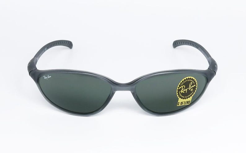 May include: Dark gray Ray-Ban sunglasses with dark green lenses. The sunglasses have a curved frame and a sticker on the right lens that reads "Ray-Ban G-15 LENS". The Ray-Ban logo is visible on the left lens.