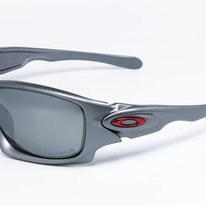 May include: Gray sunglasses with dark lenses. The sunglasses have a sleek, modern design with a red and black logo on the side. The frames are a matte gray color, and the lenses are a dark gray.