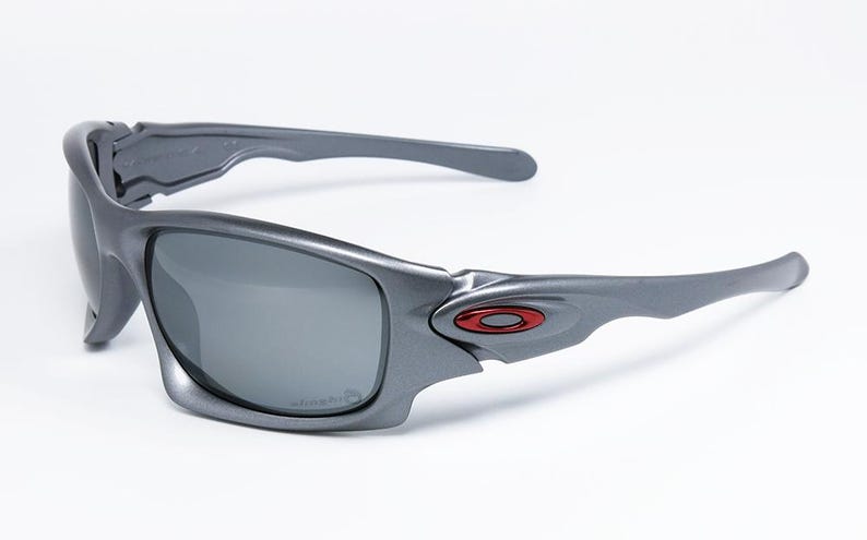 May include: Gray sunglasses with dark lenses. The sunglasses have a sleek, modern design with a red and black logo on the side. The frames are a matte gray color, and the lenses are a dark gray.