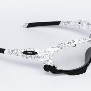 May include: White and black sunglasses with a unique design. The frames are white with a black pattern, and the arms have black tips. The lenses are clear, and the overall style is modern and sporty.