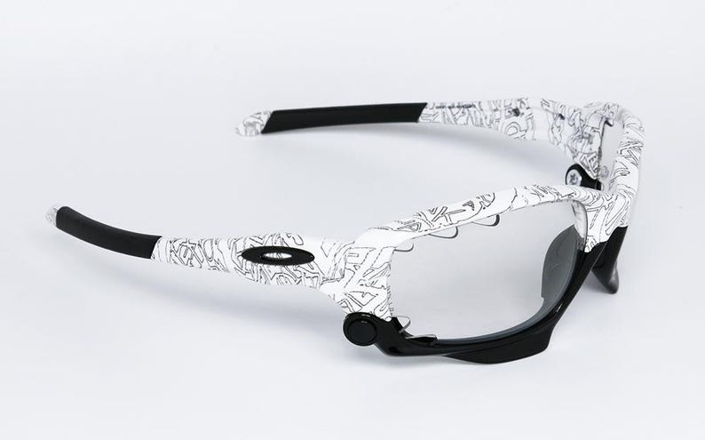 May include: White and black sunglasses with a unique design. The frames are white with a black pattern, and the arms have black tips. The lenses are clear, and the overall style is modern and sporty.