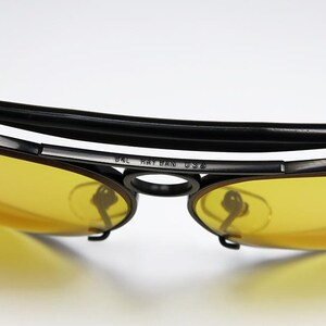 Puede incluir: Primer plano de gafas de sol vintage con lentes amarillas y montura negra. La barra superior es negra y acolchada. El texto "BAL RAY BAN USA" es visible en la montura. Las lentes son de color amarillo brillante.