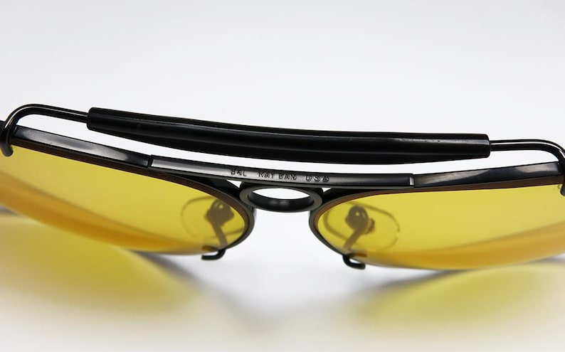 Puede incluir: Primer plano de gafas de sol vintage con lentes amarillas y montura negra. La barra superior es negra y acolchada. El texto "BAL RAY BAN USA" es visible en la montura. Las lentes son de color amarillo brillante.
