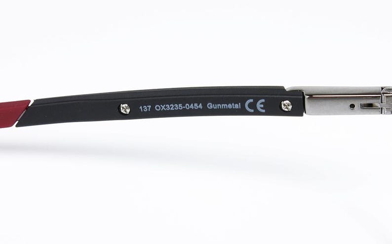 May include: Close-up of a portion of eyeglasses. The frame is black with a red accent and the text "137 OX3235-0454 Gunmetal CE" is printed on the arm. The hinge is silver.