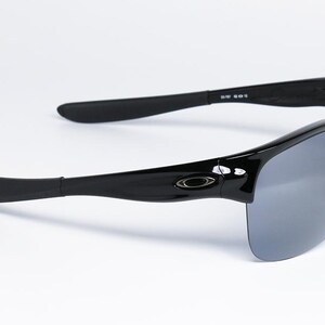 May include: Black sunglasses with gray lenses. The sunglasses have a glossy finish and a curved design. The arms of the sunglasses are thick and have a slight curve. The lenses are a gray color and are partially rimless.