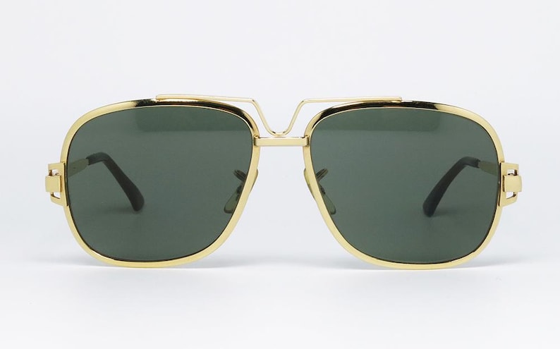 Puede incluir: Gafas de sol con montura dorada y lentes verde oscuro. Las gafas tienen un doble puente y forma rectangular. Las monturas son de color dorado brillante y las lentes son de color verde oscuro s&oacute;lido. Las gafas son de estilo aviador cl&aacute;sico.