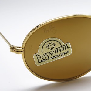 Pu&ograve; includere: Occhiali da sole dorati con lenti marroni. Il logo Ray-Ban &egrave; visibile sulla lente e sulla montatura. Un adesivo sulla lente recita "DIAMOND HARD Scratch-Protection System."