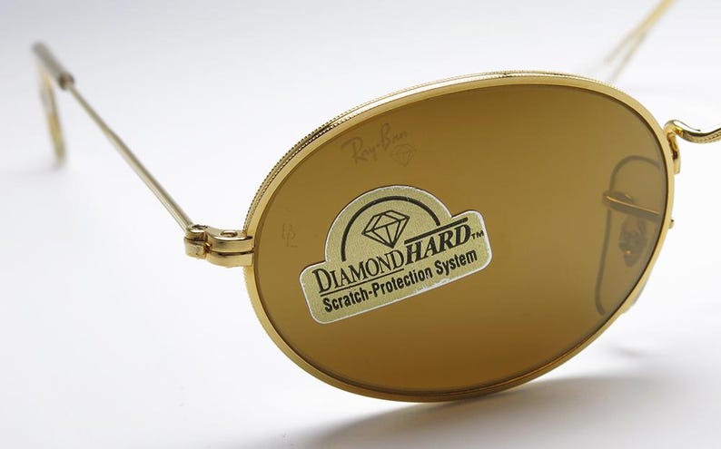 Pu&ograve; includere: Occhiali da sole dorati con lenti marroni. Il logo Ray-Ban &egrave; visibile sulla lente e sulla montatura. Un adesivo sulla lente recita "DIAMOND HARD Scratch-Protection System."