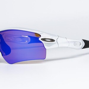 Pu&ograve; includere: Occhiali da sole sportivi bianchi e neri con lenti blu-viola. Gli occhiali hanno un design curvo con montatura bianca e aste nere. Le lenti sono di un vibrante colore blu-viola, offrendo un look sportivo e moderno.