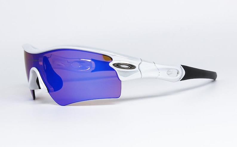 Pu&ograve; includere: Occhiali da sole sportivi bianchi e neri con lenti blu-viola. Gli occhiali hanno un design curvo con montatura bianca e aste nere. Le lenti sono di un vibrante colore blu-viola, offrendo un look sportivo e moderno.