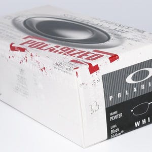 Może przedstawiać: Białe prostokątne pudełko z napisem "POLARIZED" w kolorze czerwonym. Pudełko zawiera nazwę marki "OAKLEY" i model "WHISKER". Pudełko zawiera parę okular&oacute;w z ramką z cyny i czarnymi soczewkami irydowymi.