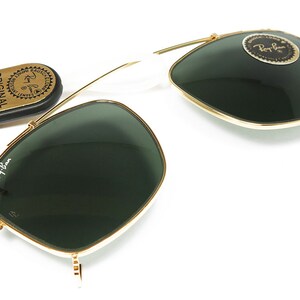 May include: Gold-rimmed sunglasses with dark green lenses and a white bridge. The Ray-Ban logo is visible on the lens and frame. A small, oval case with the text "ORIGINAL BAUSCH & LOMB" is also present.