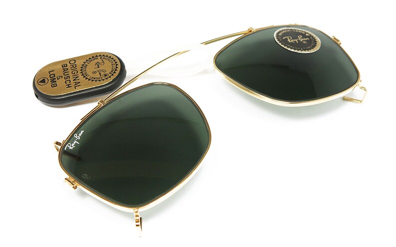 May include: Gold-rimmed sunglasses with dark green lenses and a white bridge. The Ray-Ban logo is visible on the lens and frame. A small, oval case with the text "ORIGINAL BAUSCH & LOMB" is also present.