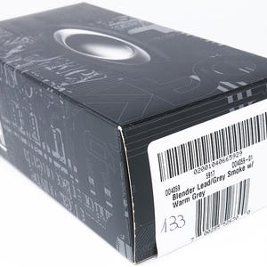 May include: A black and gray box with the Oakley logo on top. The box has a barcode and a label with the text "Blender Lead/Grey Smoke w/ Warm Grey". The box is on a white surface.