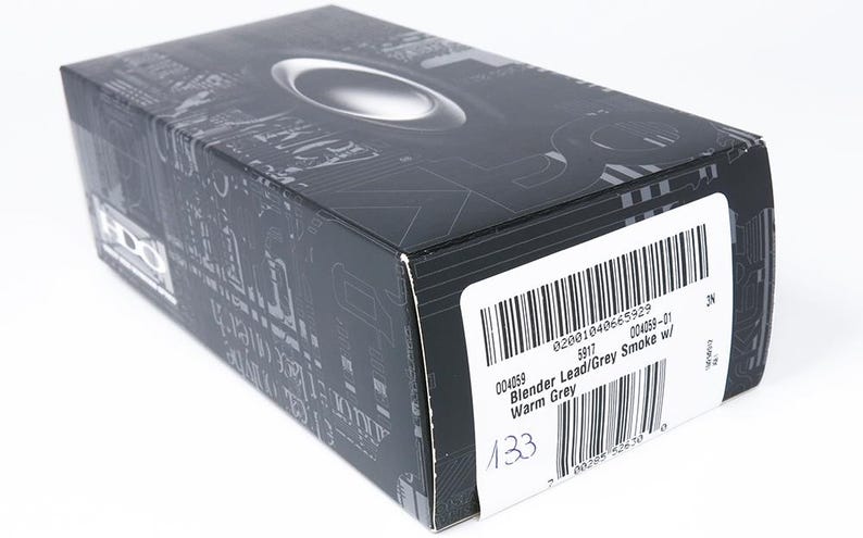 May include: A black and gray box with the Oakley logo on top. The box has a barcode and a label with the text "Blender Lead/Grey Smoke w/ Warm Grey". The box is on a white surface.