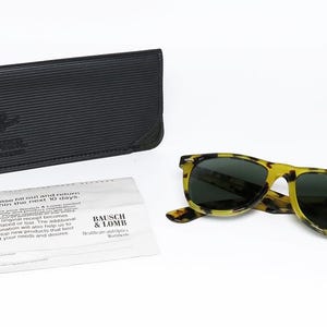 May include: A pair of yellow and black tortoise shell sunglasses with dark lenses, a black leather case, and a white paper with text. The sunglasses have a round logo on the lens.