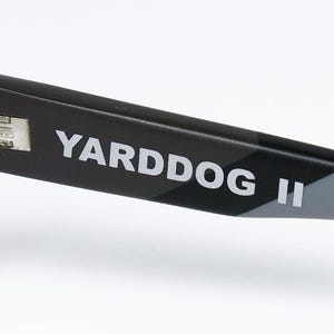 May include: Close-up of the arm of a pair of black sunglasses with the text "YARDDOG II" in white. The arm has a camouflage pattern in black and gray.